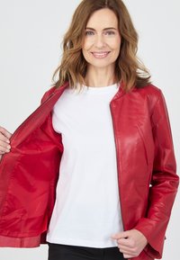 Röd läderjacka med en slät textur och dragkedja framtill, bärs över en vit t-shirt. Jackan har en figursydd design och inget synligt mönster.