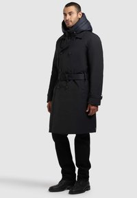 Sort trenchcoat med aftagelig quiltet hætte, dobbeltradet design, bælte i taljen og knapmanchetter. Båret over sorte bukser og støvler.
