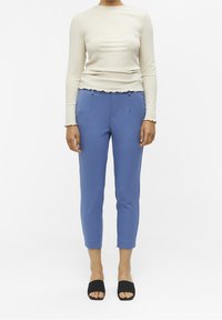 Kremowa, długosleeve'owa bluzka ze żłobieniami i falbaniastym wykończeniem, w połączeniu z jasnoniebieskimi spodniami o zwężanym kroju z przodem ze składkami oraz czarnymi klapkami.