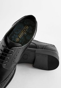 Chaussures habillées en cuir noir avec des détails de perforations, bout rond, design à lacets et semelle texturée. L'intérieur présente un revêtement à motifs floraux.