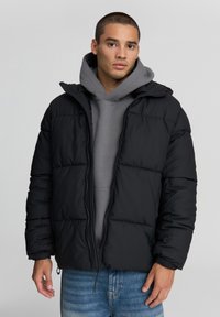 Czarna kurtka puchowa z kapturem, pikowana, z zapięciem na zamek i elastycznymi mankietami, noszona na szarym hoodie i dżinsach.