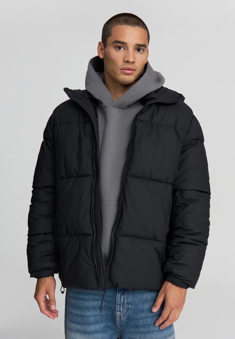 Czarna kurtka puchowa z kapturem, pikowana, z zapięciem na zamek i elastycznymi mankietami, noszona na szarym hoodie i dżinsach.