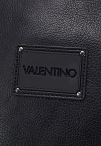 Black textured leather with a raised black logo patch and decorative silver studs. The stitching is visible around the patch.