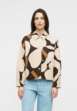 Femme aux longs cheveux bruns portant une chemise beige à boutons ornée de grandes formes abstraites noires et marron, avec un jean bleu clair, debout devant un fond blanc.