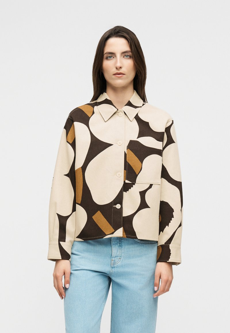 Žena s dugom smeđom kosom nosi bež košulju na kopčanje s velikim apstraktnim crnim i smeđim oblicima te svijetloplavim trapericama, stoji nasuprot bijeloj pozadini.