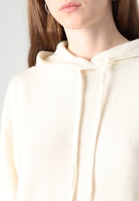 Krémová mikina s kapucňou vyrobená z jemného pleteného materiálu, s otvorom na šnúrku a voľným strihom, s jemnou štruktúrou viditeľnou na povrchu.
