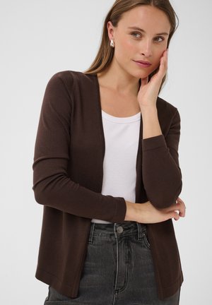 Femme portant un cardigan marron à ouverture frontale sur un haut blanc et un jean foncé, une main touchant son visage et l’autre croisée à la taille.