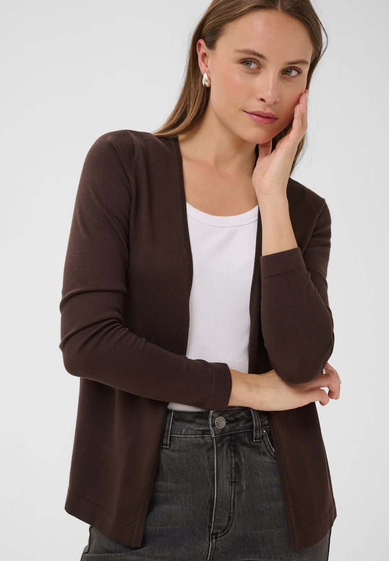 Femme portant un cardigan marron à ouverture frontale sur un haut blanc et un jean foncé, une main touchant son visage et l’autre croisée à la taille.