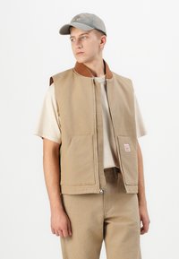 Beige canvas vest with a brown collar, featuring two front pockets and a zipper. Worn over a light-colored t-shirt, paired with beige pants.