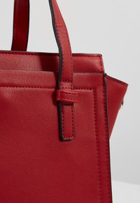 Rote Lederhandtasche mit glatter Textur, kontrastierenden schwarzen Nähten und einer strukturierten Form. Verfügt über doppelte Tragegriffe, die mit einer Schnalle befestigt sind.