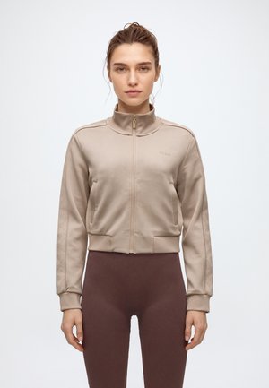 Une femme porte une veste beige zippée à col montant et aux manches à motifs, assortie à un legging marron foncé, sur un fond uni.