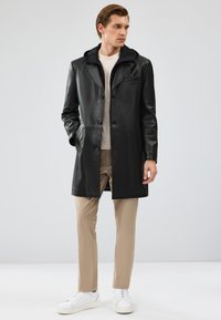 Zwarte leren jas met een textuurcapuchon, enkelvoudige knopen en zijzakken. Gedragen over een lichte top en beige broek, met witte sneakers.