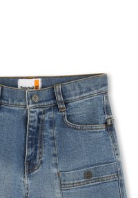 Jeans en denim avec un lavage moyen, comprenant cinq poches, une étiquette Timberland orange et blanche, et des coutures dorées contrastantes. Design classique.