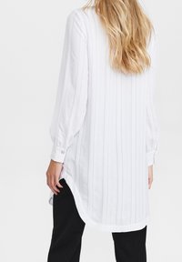 Vit randig långärmad skjorta med rundad nederkant och knäppta manschetter, tillverkad av lättviktsmaterial. I kombination med svarta byxor.