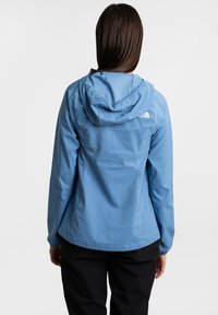 Helle blaue Windjacke mit Kapuze, die eine glatte Textur und ein dezentes Logodetail auf dem Rücken aufweist. Schwarze Hosen sind unten sichtbar.