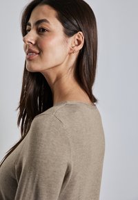 Pull en laine brun clair avec un col rond et des manches longues, présentant une texture lisse et des coutures minimales, porté par un mannequin.