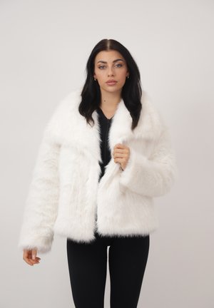 Jonge vrouw met donker haar, gekleed in een witte faux fur jas over zwarte kleding, staand tegen een effen lichte achtergrond.