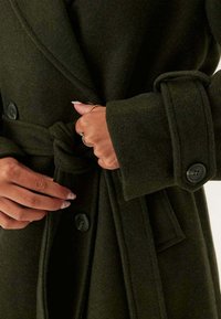 Cappotto di lana verde scuro con design avvolgente, grandi bottoni neri e dettaglio in vita legato. Il tessuto ha una finitura morbida e testurizzata.