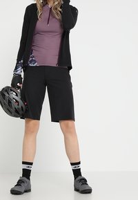 Mustat pyöräilyshortsit, joissa on purppuranvärinen vetoketjullinen paita, jossa on geometrisia kuvioita. Yhdistetty harmaisiin pyöräilykengiin ja hanskoihin. Kypärä pidetään kädessä.