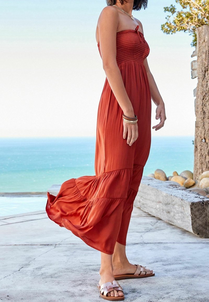 Robe maxi sans manches de couleur rouille, avec un corsage smocké, une jupe fluide et des accents froncés. Associée à des sandales plates roses. Cadre naturel en extérieur.