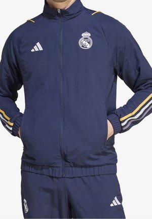 Homme portant un survêtement bleu marine du Real Madrid avec une veste zippée et un pantalon assorti, comportant les logos du club et les bandes Adidas sur les manches et les jambes.