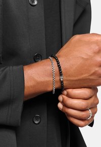 Schwarzes Perlenarmband mit kleinem Totenkopf-Anhänger, zusammen mit einem silbernen Kettenarmband, getragen an einer Hand. Dunkle Kleidung im Hintergrund.