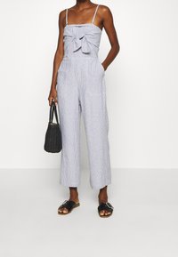Vrouw in een lichtblauwe en witte gestreepte mouwloze jumpsuit met een strik aan de voorkant, die een zwarte gevlochten handtas draagt en zwarte slide sandalen met een gesp.