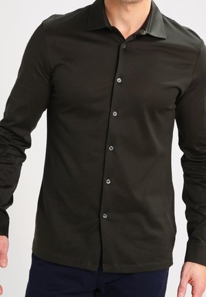 Mann in einem eng anliegenden, schwarzen Langarmhemd mit klassischem Kragen und dunkelblauer Hose vor einem schlichten Hintergrund.