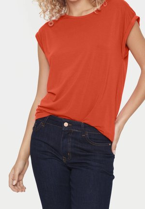 Kobieta ubrana w jasnopomarańczowy T-shirt z krótkim rękawem, wsunięty w ciemnoniebieskie dżinsy, ręka spoczywa w tylnej kieszeni.