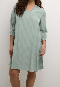 Helles grünes Kleid mit V-Ausschnitt und Dreiviertelärmel, das ein strukturiertes karriertes Muster und eine lockere Passform aufweist.