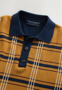 Stribet polo skjorte i sennep gul med en marinblå krave og tre sorte knapper. Udsmykket med hvide og marinblå vandrette linjer.