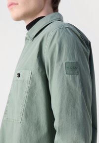 Homme portant une chemise légère verte à boutons avec une poche poitrine et un écusson logo "BOSS" sur la manche supérieure, sur un fond blanc.