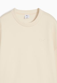 Camiseta de color beige claro hecha de una tela suave, con un cuello redondo y mangas cortas, y con un pequeño logo bordado en la parte frontal.