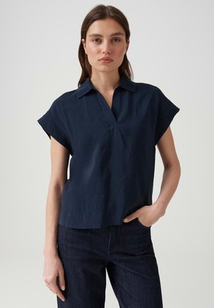 Femme debout avec une main dans la poche, portant un chemisier bleu marine à manches courtes avec col et un jean foncé sur un fond clair uni.