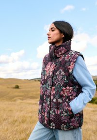 Steppweste mit Blumenmuster in dunkelviolett, mit hohem Kragen und zwei Fronttaschen, kombiniert mit einem hellblauen Langarmshirt.