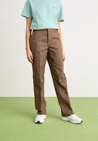 Pantalons ajustés marron avec une taille plate et des poches latérales, associés à un t-shirt en coton bleu clair. Portés avec des baskets blanches.