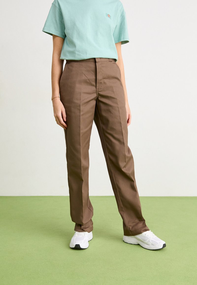 Pantalons ajustés marron avec une taille plate et des poches latérales, associés à un t-shirt en coton bleu clair. Portés avec des baskets blanches.