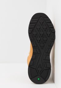 Semelle en caoutchouc noire avec des crampons en forme de croix et un petit hexagone vert, attachée au côté d'une botte de randonnée beige sur fond blanc.
