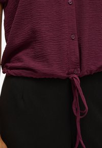 Maroon, textured blouse with a button front and gathered waist, featuring a tie detail. Paired with black trousers, textured fabric visible.