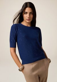 Donna con lunghi capelli scuri che indossa un maglione blu navy a maniche corte con piccole macchie bianche e pantaloni beige a vita alta con le mani nelle tasche.