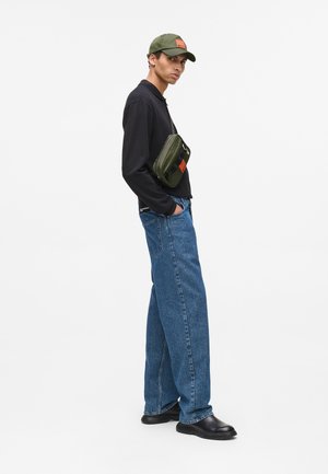 Model trägt ein schwarzes Langarmshirt, lockere blaue Jeans, schwarze Schuhe und eine olivgrüne Mütze mit einem rechteckigen orangefarbenen Patch.