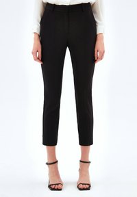 Vrouw in zwarte slim-fit enkellange broek, witte blouse en zwarte open teen hakken die tegen een effen achtergrond staat.