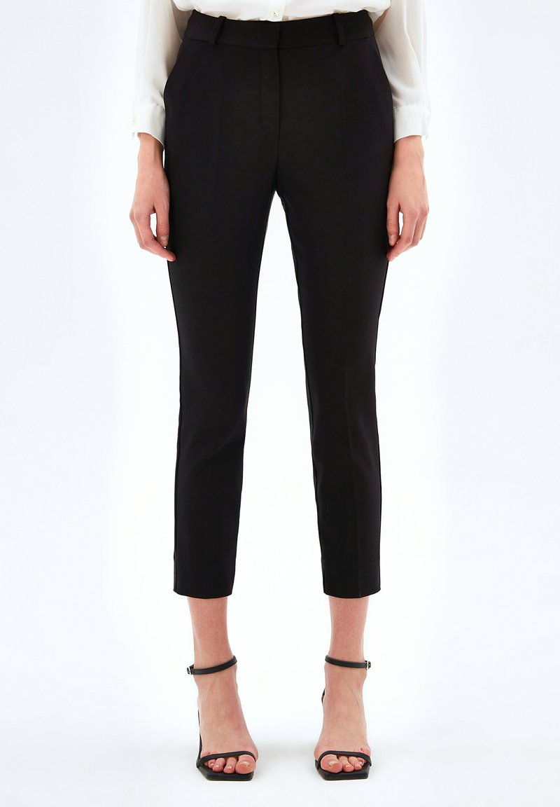 Vrouw in zwarte slim-fit enkellange broek, witte blouse en zwarte open teen hakken die tegen een effen achtergrond staat.