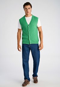 Grön stickad väst med v-ringning, med kabelmönster och knappar framtill, bärs över en vit t-shirt och kombineras med blå jeans.