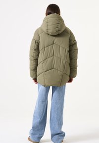 Chaqueta acolchada de color verde oliva con capucha, que presenta patrones en diagonal acolchados y un corte relajado, combinada con jeans de pierna ancha de color azul claro.