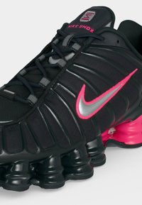 Μαύρο sneaker Nike Shox με ροζ και ασημένιο λογότυπο Swoosh, μαύρα κορδόνια και χαρακτηριστική ελαστική απορρόφηση κραδασμών σε μαύρο και ροζ χρώμα.