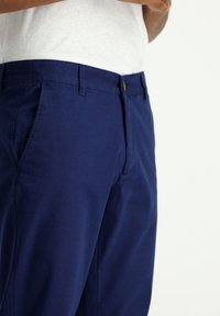 Pantaloni blu navy in tessuto resistente, con chiusura frontale a bottone, passanti per la cintura e tasche laterali. Texture liscia e vestibilità tailored.
