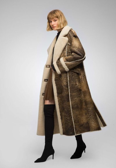 Shearling coat with brown and cream tones, featuring a wide collar, toggle closures, and contrasting textures. Model wearing knee-high boots.