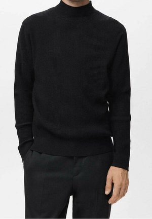 Homme portant un pull en maille côtelée noir à col montant simulé et un pantalon noir, debout avec une main dans la poche et l'autre détendue sur le côté.