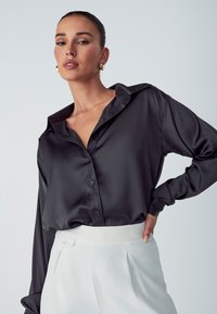 Camicia nera in satin con colletto, maniche lunghe e chiusura con bottoni, abbinata a pantaloni bianchi a vita alta.